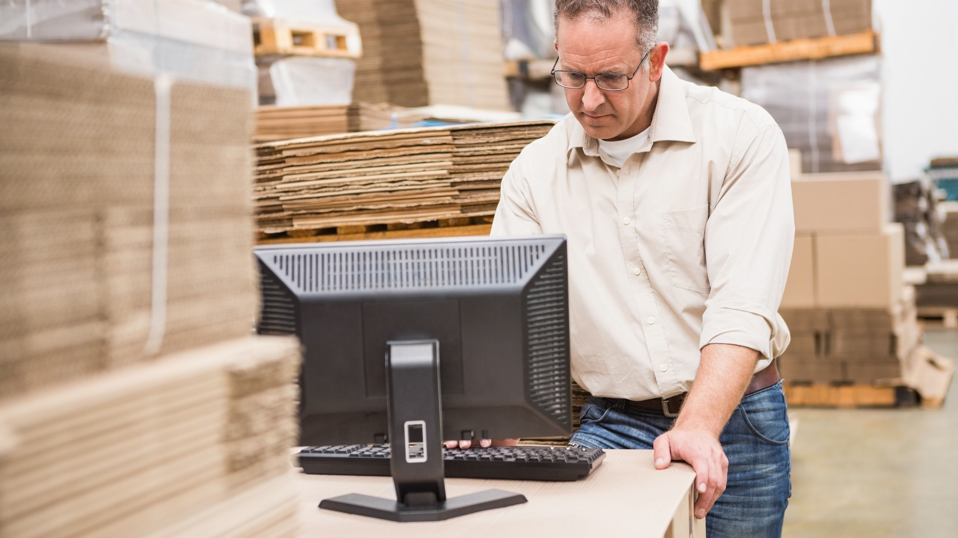 Empleado de almacén consultando el sistema en un monitor de sobremesa junto a palés y cajas